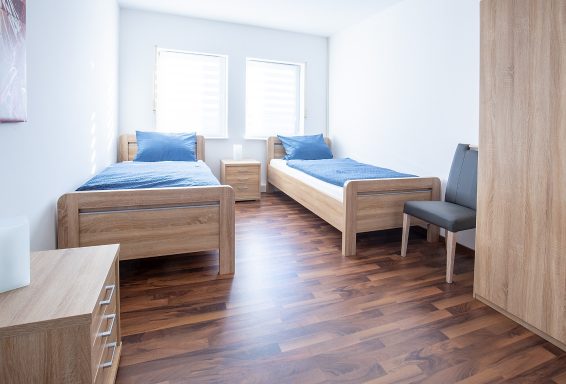 Helles Zimmer mit zwei Einzelbetten, Nachttisch und einem Stuhl. Holzfußboden.