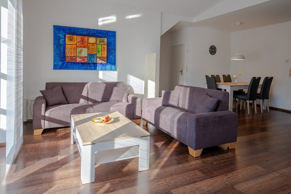 Helle Wohnraum mit zwei lila Sofas, einem Tisch und einem Esstisch im Hintergrund.