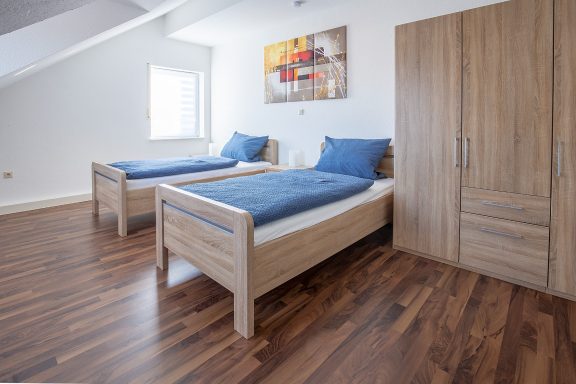 Zimmer mit zwei Einzelbetten, Holzfußboden und einem Schrank, hell und modern eingerichtet.