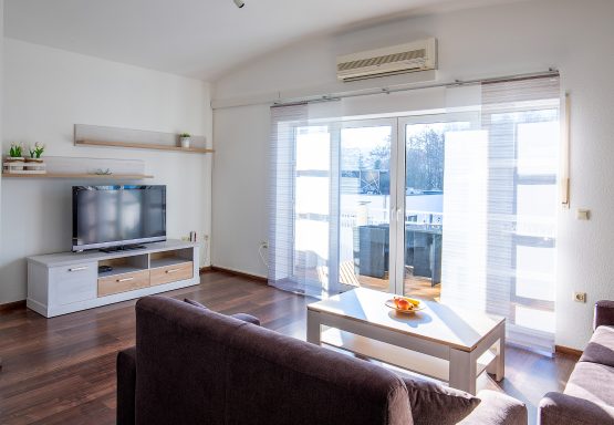 Helles Wohnzimmer mit Fernseher, Sofa und Balkonzugang.
