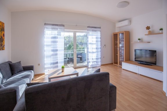 Heller Wohnraum mit grauem Sofa, Glastisch, TV und Balkonzugang.